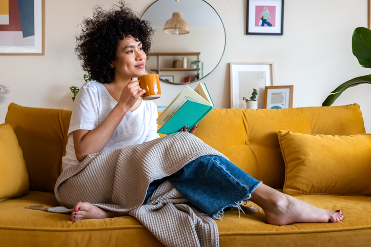 Pensive relaxed African american woman reading a book at home, drinking coffee sitting on the couch.