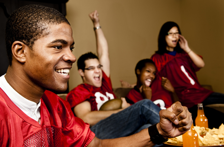 Multi-ethnic group of football fans, friends at home watching the game on television. They are cheering. Snacks.